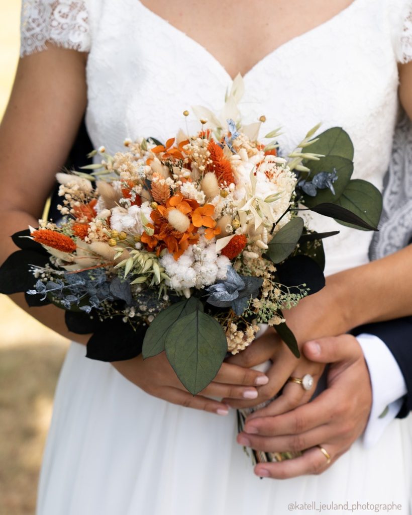 Mariée en robe blanche tenant un bouquet de fleurs séchées terracotta, blanches et feuillage vert foncé - Les pensées de Lily