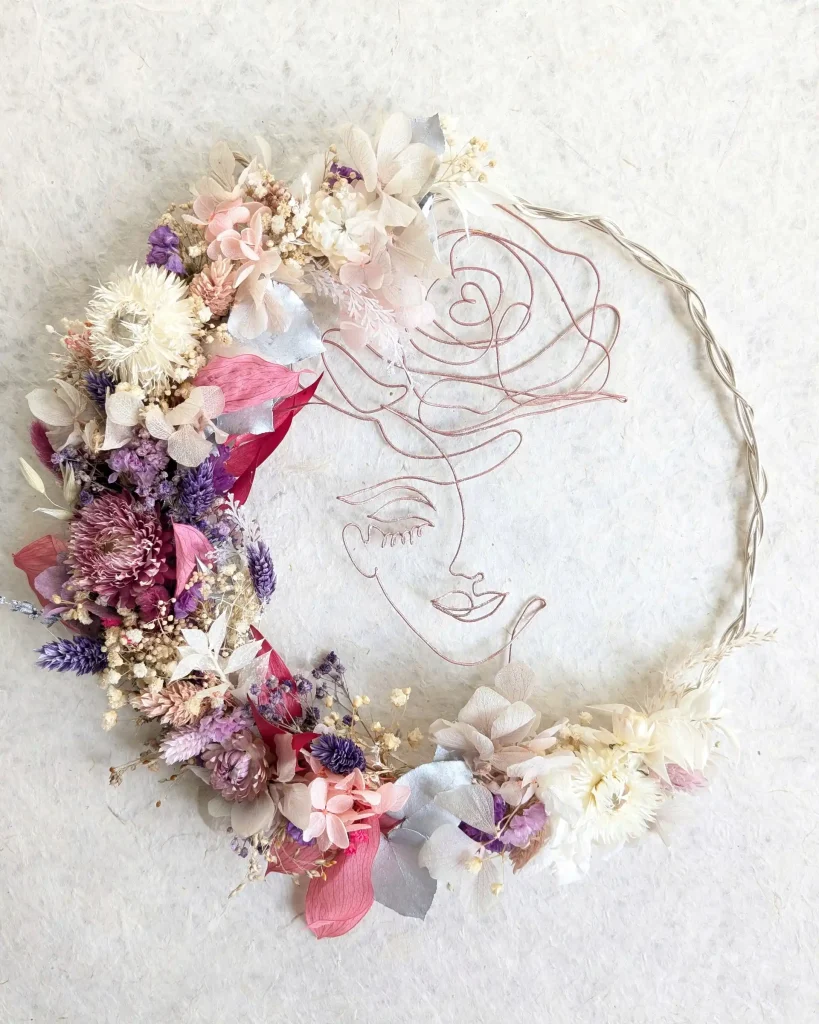 Couronne murale en fleurs séchées aux tons mauve, rose, crème et bordeaux, avec un visage de femme et une rose dessinés en fil rose doré – Les pensées de Lily
