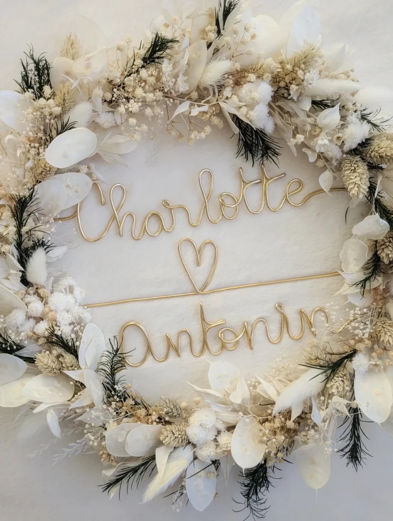 Couronne de mariage en fleurs séchées blanches et feuillage vert sombre, décorée de deux prénoms en fil doré reliés par un cœur, conçue pour une décoration personnalisée de cérémonie ou de réception – Les pensées de Lily