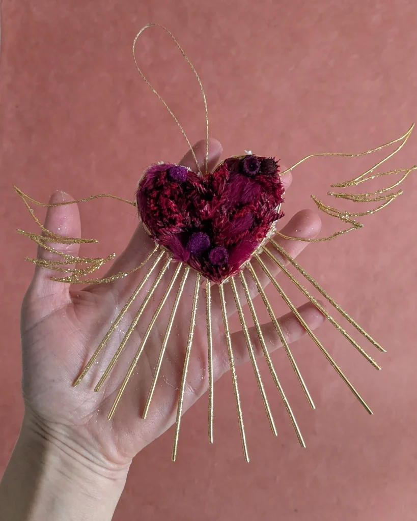 Ex-voto suspendu en cœur de fleurs séchées rouges et ailes dorées, tenu en main devant un fond rose – Les pensées de Lily