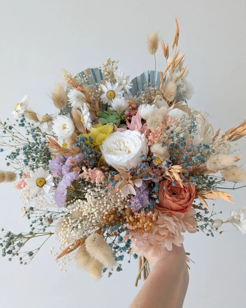 Bouquet coloré en fleurs séchées pastel avec des immortelles blanches, roses saumon, gypsophile bleue et blé blond, vu de face, tenu à la main - Les pensées de Lily.