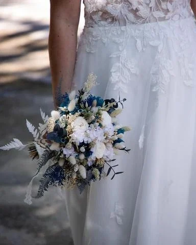 Bouquet de mariée en fleurs séchées aux tons bleu nuit, ivoire et vert fougère, tenu à la main avec une robe blanche en dentelle – Les pensées de Lily.