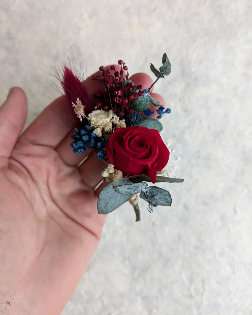 Boutonnière en fleurs séchées rouge et bleu avec eucalyptus, présentée en main - Les pensées de Lily