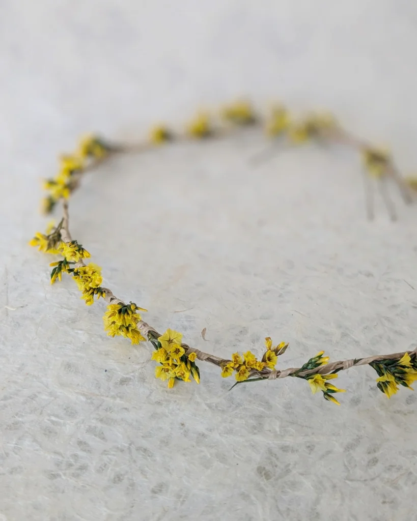 Couronne fine en fleurs séchées jaunes et feuillage vert, disposée à plat sur fond texturé clair – Les pensées de Lily