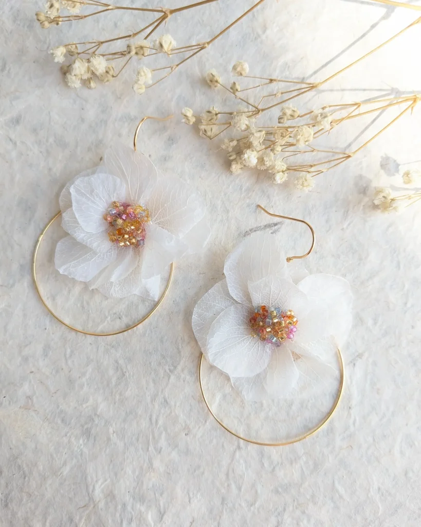 Paire de boucles d’oreilles en fleurs séchées blanches avec cœur de perles orangées, rosées et dorées, posées sur papier naturel – Les pensées de Lily