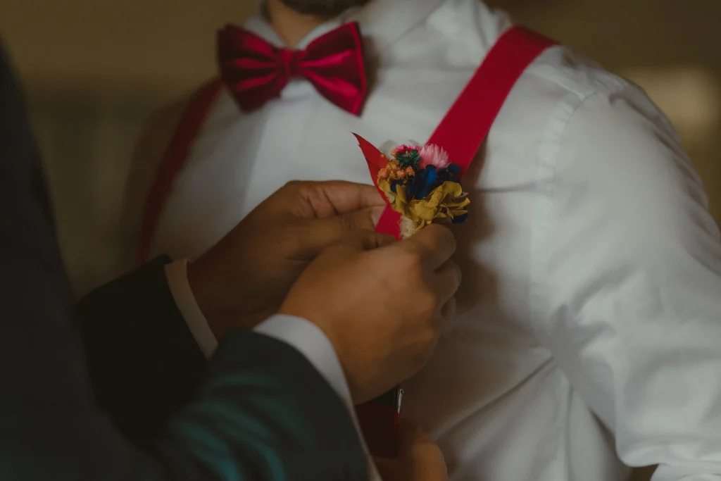 Main ajustant une boutonnière en fleurs séchées colorées sur la bretelle rouge du témoin - Les pensées de Lily