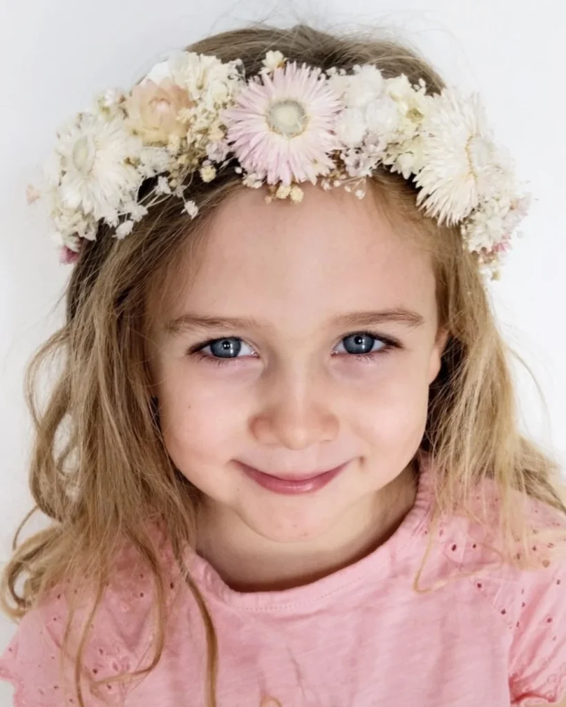 Petite fille blonde souriante portant une couronne de fleurs séchées blanc et rose sur fond clair - Les pensées de Lily