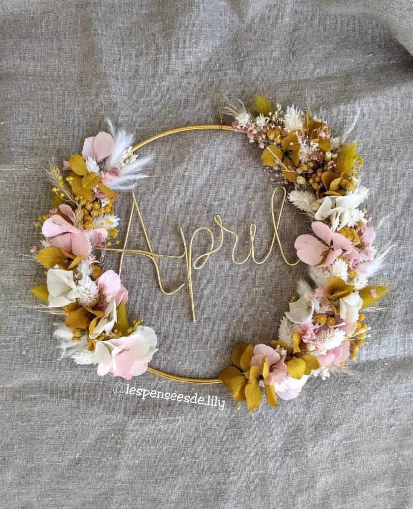 Couronne prénom April en fil doré, décorée de fleurs séchées roses, blanches, jaune moutarde et feuillage sur fond en lin naturel - Les pensées de Lily