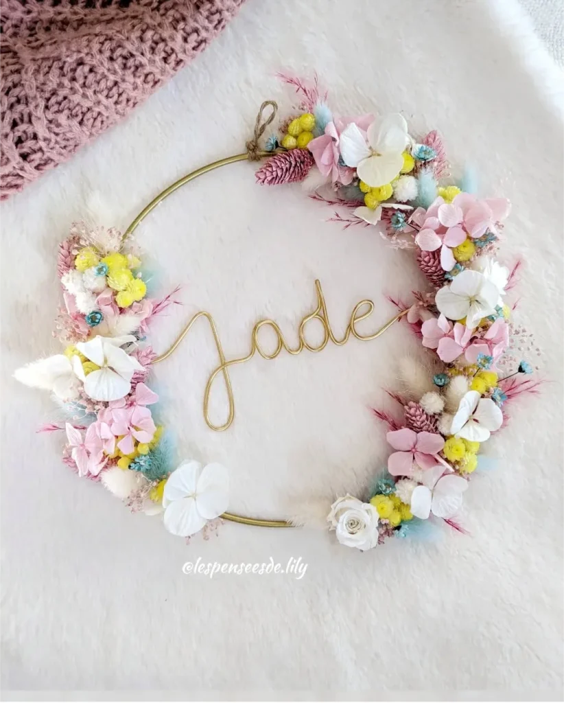 Couronne murale avec le prénom Jade en fil doré, décorée de fleurs séchées pastel, blanches, roses, jaunes et touches de bleu sur fond doux - Les pensées de Lily