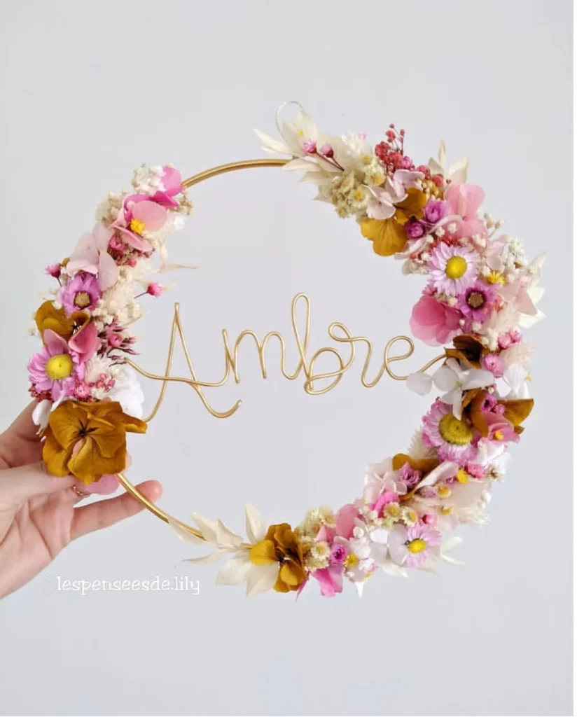 Couronne murale avec le prénom Ambre en fil doré, décorée de fleurs séchées blanches, roses et jaune moutarde tenue à la main - Les pensées de Lily