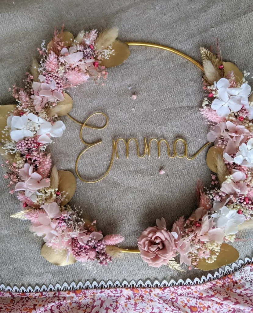 Couronne murale avec le prénom Emma en fil doré, ornée de fleurs séchées blanches, rose poudré, feuillages dorés et rose centrale - Les pensées de Lily