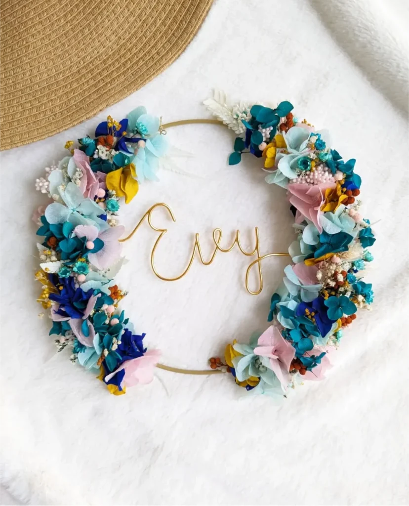 Couronne murale avec le prénom Evy en fil doré, décorée de fleurs séchées bleu roi, turquoise, rose pastel et touches de jaune sur fond blanc - Les pensées de Lily