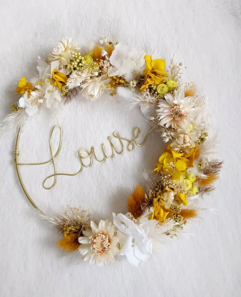 Couronne murale avec le prénom Louise en fil doré, décorée de fleurs séchées jaunes, blanches, crème et éléments naturels sur fond blanc - Les pensées de Lily