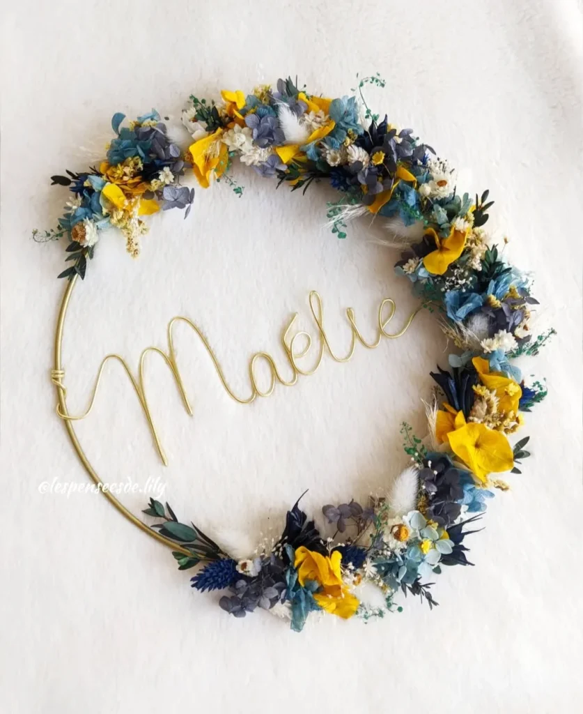 Couronne murale avec le prénom Maélie en fil doré, décorée de fleurs séchées bleues, jaunes, blanches et feuillage vert sur fond blanc - Les pensées de Lily