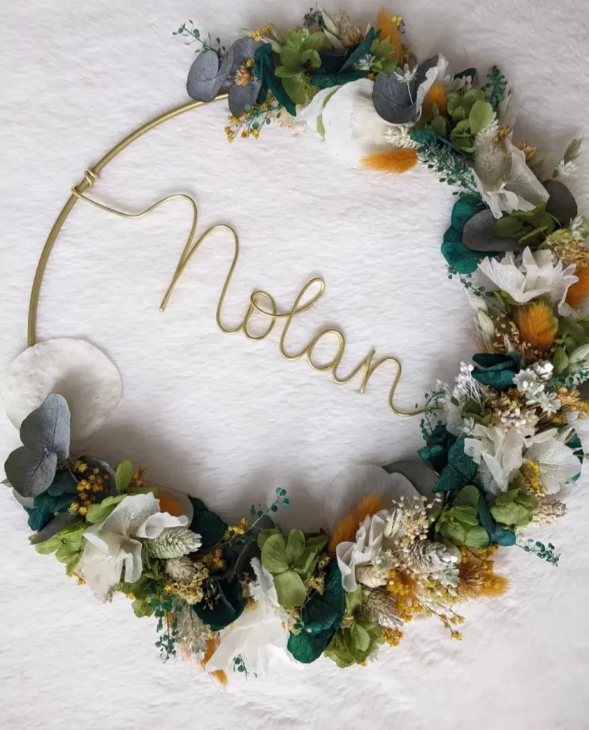 Couronne murale personnalisée avec le prénom Nolan en fil doré, décorée de fleurs séchées blanches, feuillage vert, touches de jaune et eucalyptus sur fond clair - Les pensées de Lily