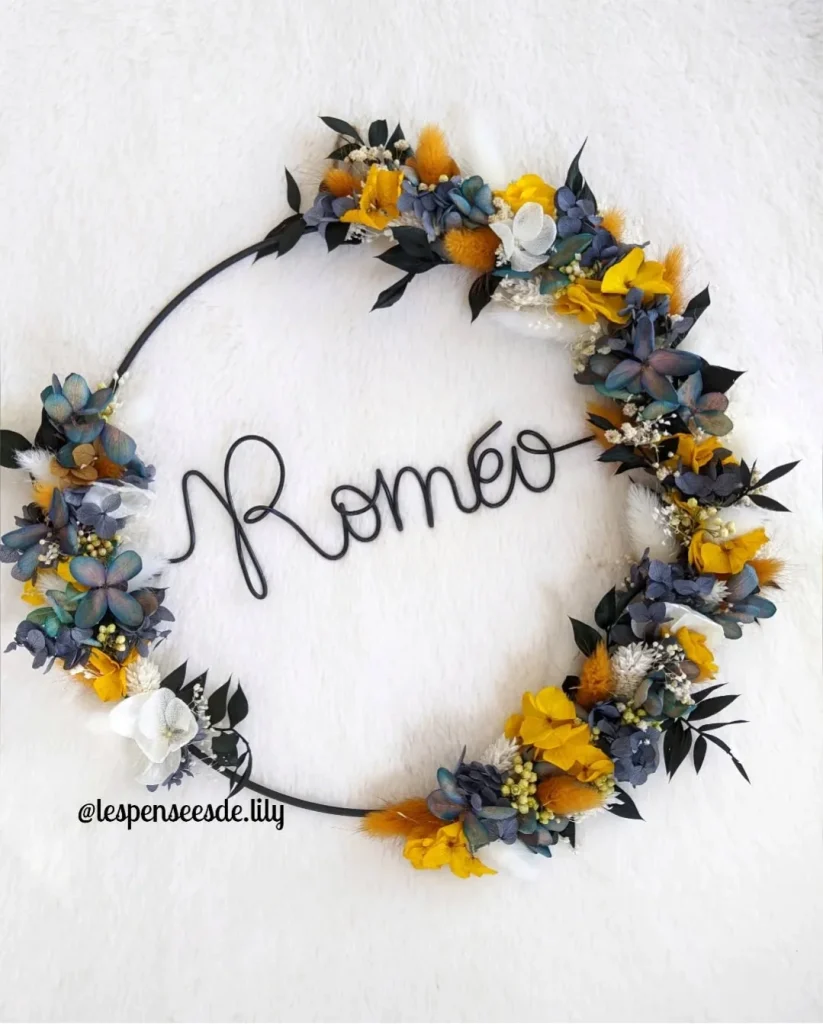 Couronne murale avec le prénom Roméo en fil noir, ornée de fleurs séchées bleu-gris, jaunes vives, blanches et feuillage foncé sur fond blanc - Les pensées de Lily