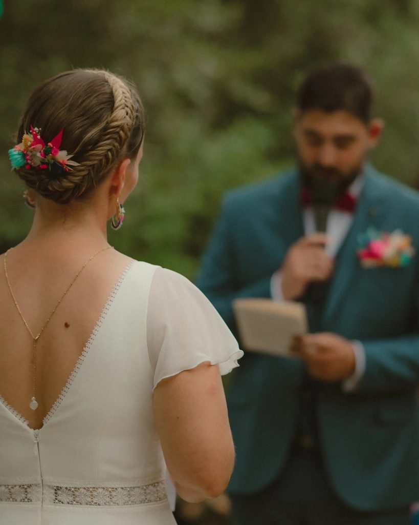 Mariée de dos avec coiffure tressée fleurie écoutant le marié lire ses vœux dans un décor naturel - Les pensées de Lily